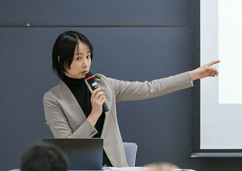 マイクを持ち、スクリーンを指し示しながら説明する女性講師